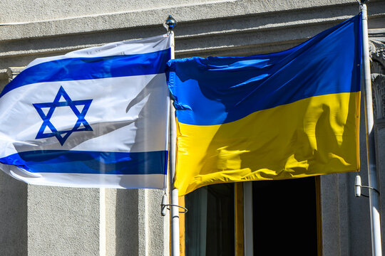 The national flags of Ukraine and Israel wave on flagpoles near an administrative building under bright sunlight, symbolizing diplomatic ties, political cooperation, and mutual support.