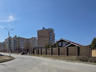 Obraz premium Modern residential apartment buildings and a private house with a tall wooden fence along a city street under a clear blue sky