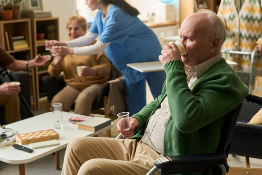 Senior man in wheelchair drinking from cup during group care session while nurse serving water to elderly patients, supporting assisted living, caregiving marketing