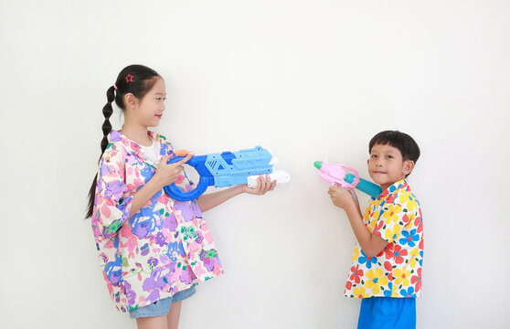 Two children, a girl and a boy wearing colorful floral shirts, are having fun playing with water guns against white background, Ready for Songkran festival celebrations.