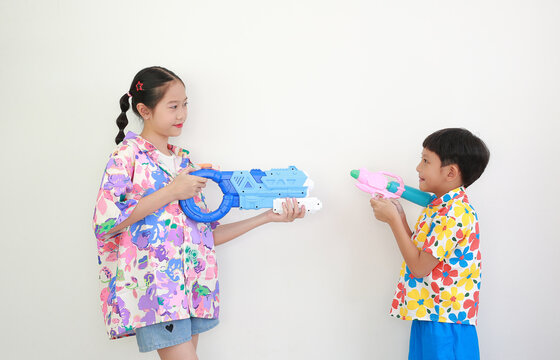 Two children, a girl and a boy wearing colorful floral shirts, are having fun playing with water guns against white background, Ready for Songkran festival celebrations.
