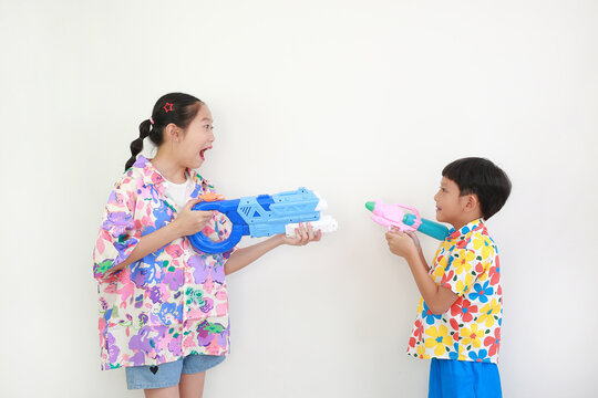 Two children, a girl and a boy wearing colorful floral shirts, are having fun playing with water guns against white background, Ready for Songkran festival celebrations.