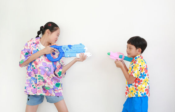 Two children, a girl and a boy wearing colorful floral shirts, are having fun playing with water guns against white background, Ready for Songkran festival celebrations.