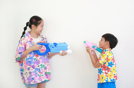Two children, a girl and a boy wearing colorful floral shirts, are having fun playing with water guns against white background, Ready for Songkran festival celebrations.