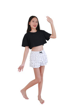 Cheerful Asian preteen girl in black crop top and polka-dot ruffled shorts posing playfully and waving her hand, isolated on white background.