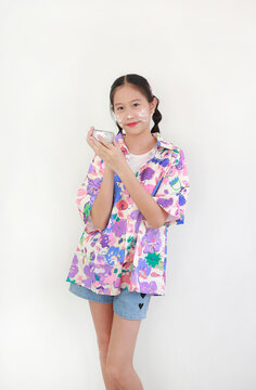 Smiling young Asian girl in colorful floral shirt celebrates Songkran Festival, holding traditional silver water bowl with white clay powder (Din Sor Pong) smeared on her cheeks on white background.