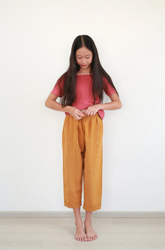 An adorable Asian girl with long hair wearing a pink t-shirt and yellow trousers, standing barefoot while focusing on tying her waistband against white wall.