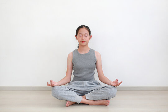 A young Asian girl sitting in lotus position with her eyes closed, practicing meditation and mindfulness in a bright, minimalist room.