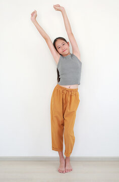 Cheerful young girl with long dark hair in grey sleeveless top and mustard-colored trousers, stretching her arms high above her head with a happy expression against white wall.