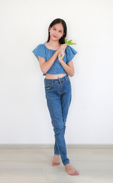 Portrait of lovely young Asian girl with long dark hair, dressed in blue crop top and denim jeans, standing barefoot and holding a small bouquet of colorful flowers while smiling in white room.