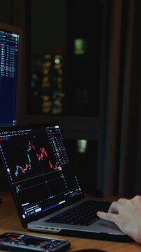 Man stock trader in home office. Male analyzes terminal, trades stocks on crypto market. Smartphone monitor and laptop display vivid candlestick chart, trading charts, price action. Evening session