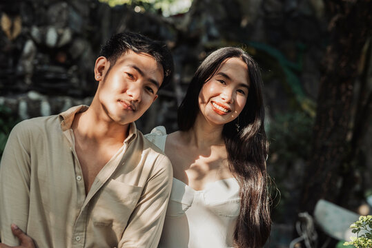 A cheerful young Asian couple smiling together under tree shade enjoying a warm sunny afternoon moment. Outdoor shot.