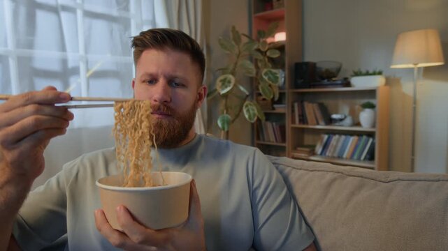 Chest up shot of young Caucasian man in casualwear sitting on couch and slurping instant noodles using chopsticks, enjoying convenience food at home