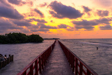Obraz premium Perspective of Red Wooden Bridge over the Sea at Golden Sunset,A long red wooden bridge stretches into the sea towards the horizon under a dramatic golden sky during sunset. The scene features lush.