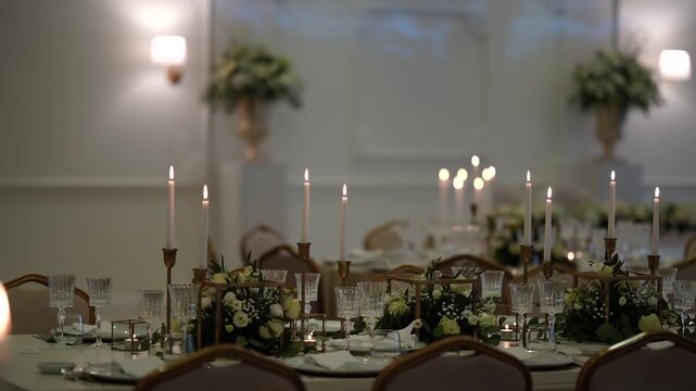 Long banquet dining table with gold candelabras, lit taper candles, white floral garland, crystal glassware and place settings in white ballroom