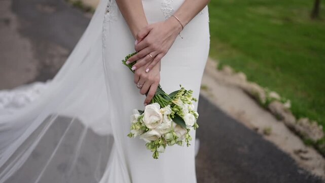 Bride in white lace gown holding white rose bouquet with both hands, bracelet and ring visible, green grass and path in background