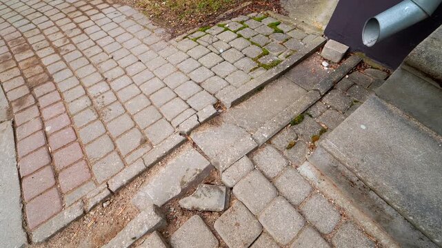 Uneven broken paving stones and cracked curb create a trip hazard near a building entrance and drainpipe, showing poor pavement repair and urban wear.