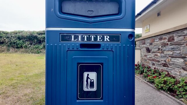 Blue outdoor litter bin near a brick building, grassy area, and landscaped shrubs.