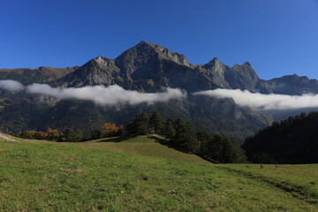 Fototapeta premium Misty peaks rise above green meadows. Clouds drift between rugged mountain ridges. Clear blue sky frames the tranquil scene. Nature's grandeur invites calm reflection