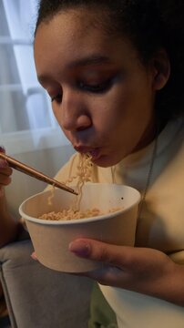 Vertical handheld arc shot of young Black girl slurping instant noodles using chopsticks from takeaway bowl at home in evening