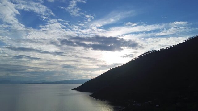 Cinematic drone shot slowly moving to the left, capturing the soft silhouette of morning light over Lake Toba