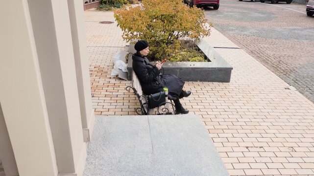Elegant woman in black beret and coat sitting on a city bench using her smartphone