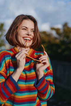 Woman smiling in a colorful striped sweater holding sunglasses outdoors; young woman portrait with bob haircut, casual outfit and joyful mood, lifestyle fashion shot in warm sunlight.