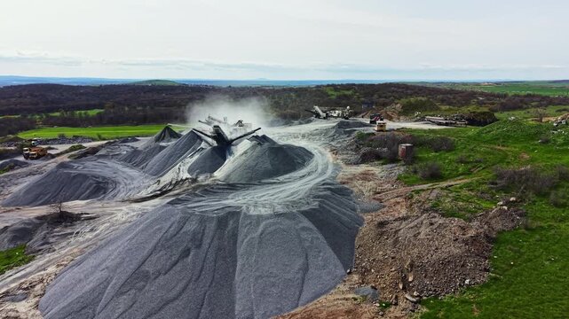 Granite is being extracted in the quarry with machines. Large hills of gravel sit around the site. Dust rises from the equipment. Green fields are in the background under a blue sky.