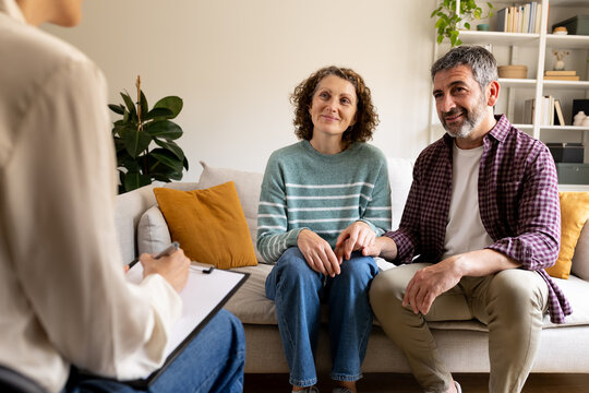 Couple sitting on a couch, holding hands, talking to a therapist writing notes