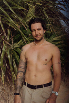 Shirtless tattooed man smiling and winking at camera by pandanus
