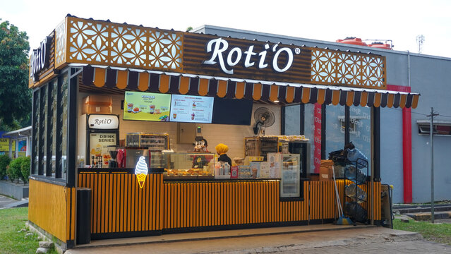 A modern exterior view of a Roti'O outlet located at a rest area along the Trans-Java Toll Road. The kiosk features its signature yellow and brown wooden slat design, decorative white floral patterns.
