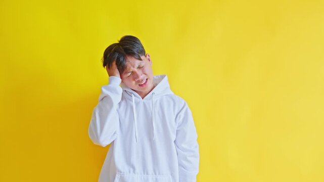 Teenage boy in hoodie clutching head with painful expression suffering from severe headache or migraine against yellow background