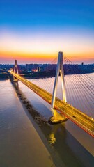 Obraz premium A stunning aerial view captures a modern cable-stayed bridge illuminated at dusk, stretching across a wide river with a vibrant cityscape visible in the background.