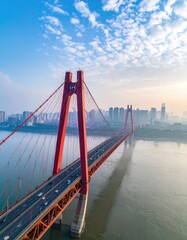 Fototapeta premium A stunning aerial view showcases a modern red suspension bridge spanning a wide river, with a sprawling cityscape bathed in the soft light of dawn.