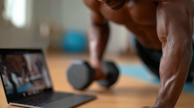 Muscular man performing renegade row exercise with dumbbell near laptop at home