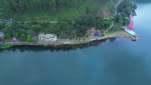 
Aerial drone view of a quiet lakeside area bordered by dense green forest and hillside roads.