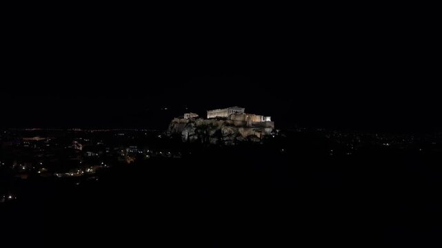 Drone footage approaches the illuminated sacred rock of the Acropolis from complete darkness, gradually revealing the Parthenon and the historic monument above Athens at night.