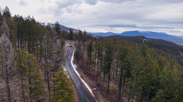 Aerial drone shot following a narrow winding mountain road cutting through evergreen and deciduous trees, with snow patches and distant mountains under a cloudy sky near Whitefish, Montana.
