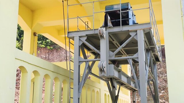 Side view shows industrial gantry and crane mechanism for dam gates. Steel structure stands on rails near yellow concrete wall. Heavy industrial gantry supports the crane mechanism.