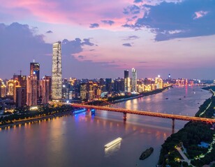 Fototapeta premium A stunning aerial view captures a vibrant cityscape illuminated at dusk, showcasing modern skyscrapers, a winding river, and a gracefully arched bridge under a colorful sky.