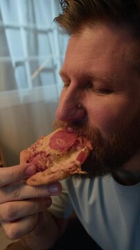 Vertical slowmo closeup shot of bearded young Caucasian man enjoying slice of delicious pizza at home