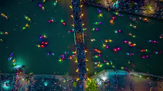 Aerial hyperlapse of illuminated wooden boats and a crowded pedestrian bridge over the river at night in Hoi An, Vietnam.