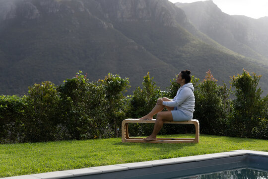 Wooden bench is sitting beside pool on trimmed lawn, hedge shielding view and mountains rising
