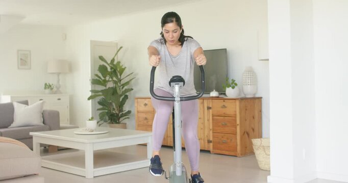 Mid adult woman gripping handlebars and pedaling on stationary bike in home living room, training