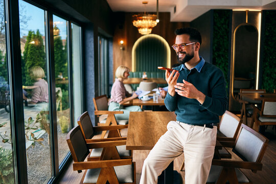 Young adult man using smartphone to record voice message while standing in modern cafe interior