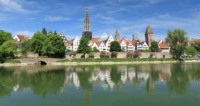 14.05.2025. View across the Danube of the Ulm old town with the Minster and Metzgerturm reflecting in the water. people cycling and relaxing by the river under a sunny blue sky. Ulm, Baden-Wuerttember