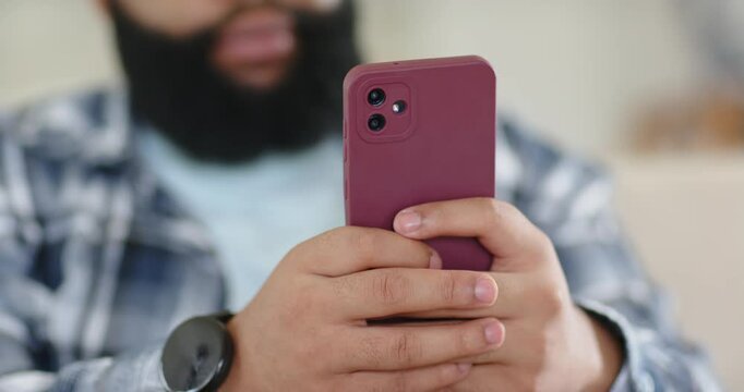 Adult man on couch bringing maroon phone forward, holding two-handed, scrolling feed, watch visible