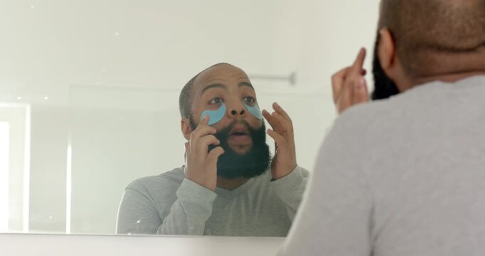 Inspecting adult man applying blue under-eye patches in bathroom mirror, wearing light grey top
