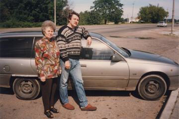 Naklejka premium Mother and son standing together next to silver hatchback car on roadside on sunny day, analog color film mid 1990s
