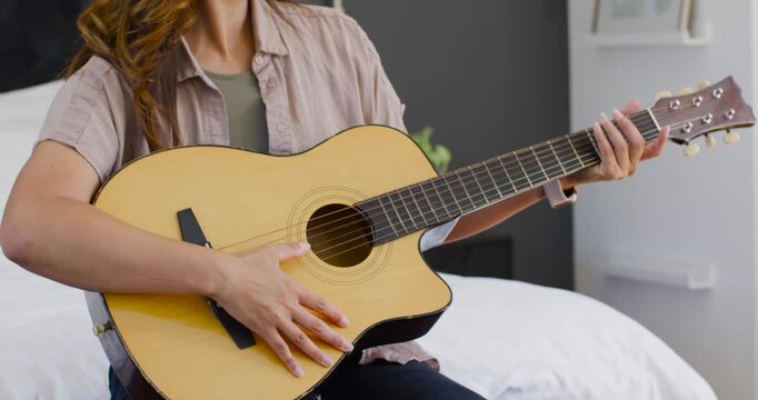 African American woman playing acoustic guitar on bed, glancing toward phone then checking it